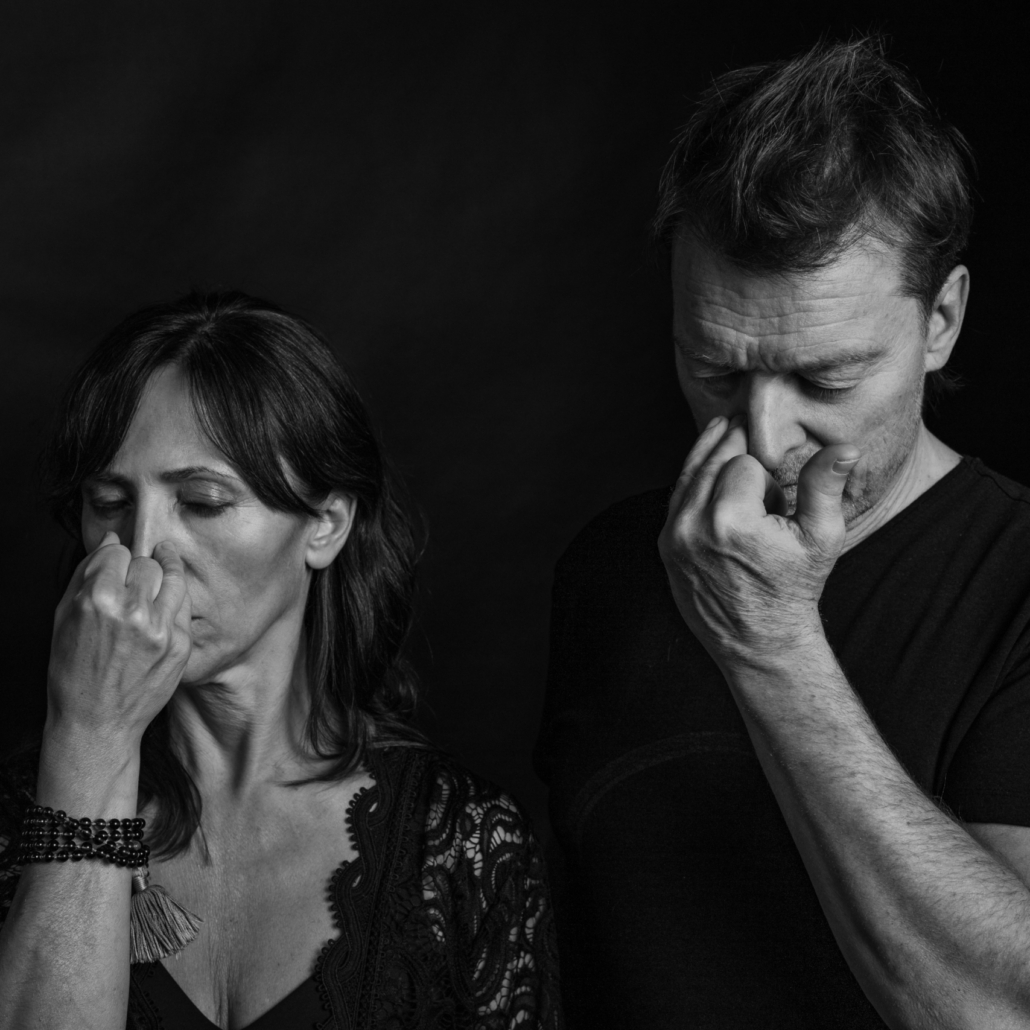 The-Bunerus-by-Ingo-Bollhoefer-MedRes-4929 Die Atemtreiner Michaela und Jörg Buneru stehen, schwarz gekleidet vor schwarzem Hintergrund, mit gesclossenen Augen nebeneinander. Sie haben eine Hand an ihrem Nasenflügel und üben konzentriert Breathwork undAtemübungen