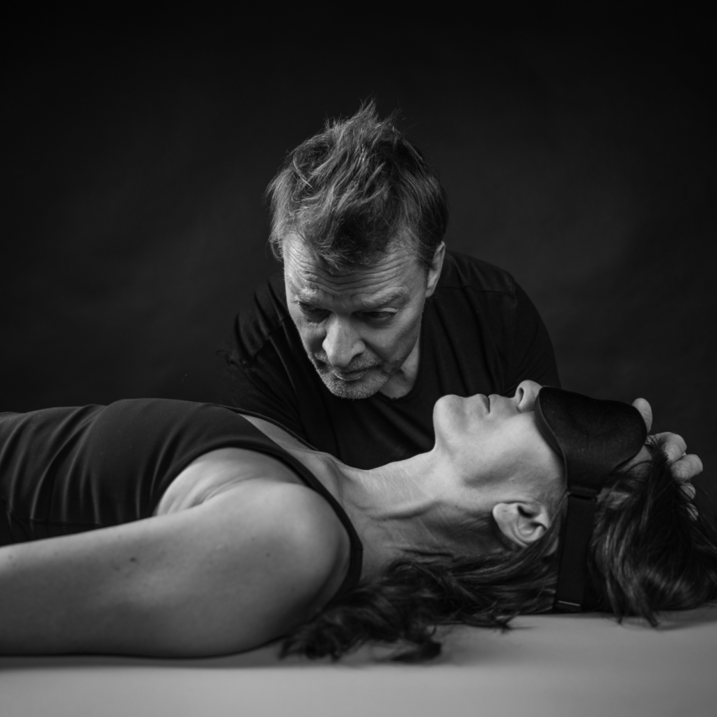 Paar Hände Herz Breathwork Trainer Michaela und Jörg Buneru stehen in einem dunkeln Raum ganz nah beieinander. Sie lächeln und vormen jeweils mit einer Hand gemeinsam ein Herz. Ihre Hände sind dabei im Fokus und im Vordergrund