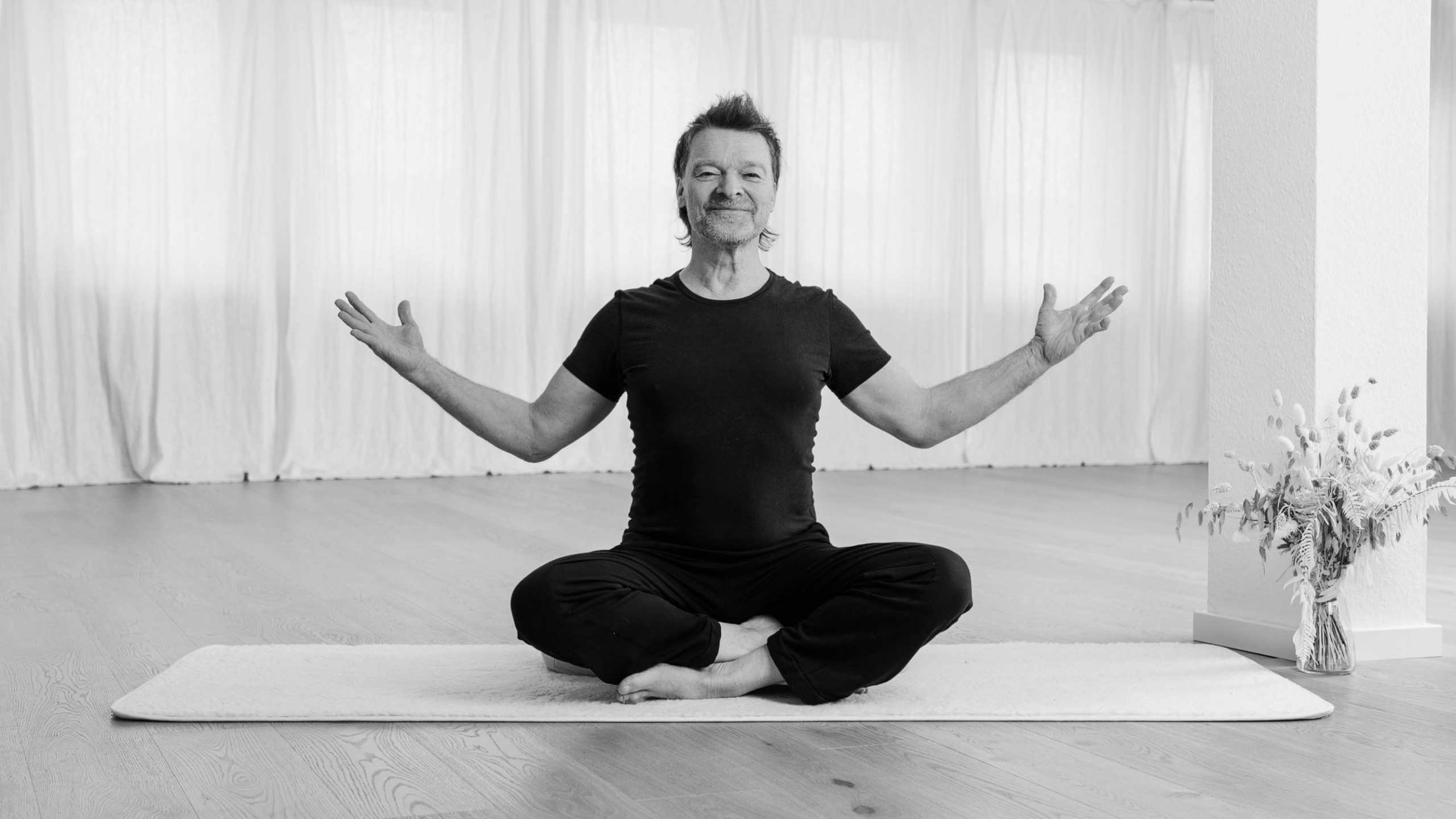 Pranayama und Breathwork Lehrer Jörg Buneru sitzt auf einer Yogamatte im Schneidersitz in einem hellen Yogastudio. Er blickt lächelnd in die Kamera, die Arme einladend geöffnet.